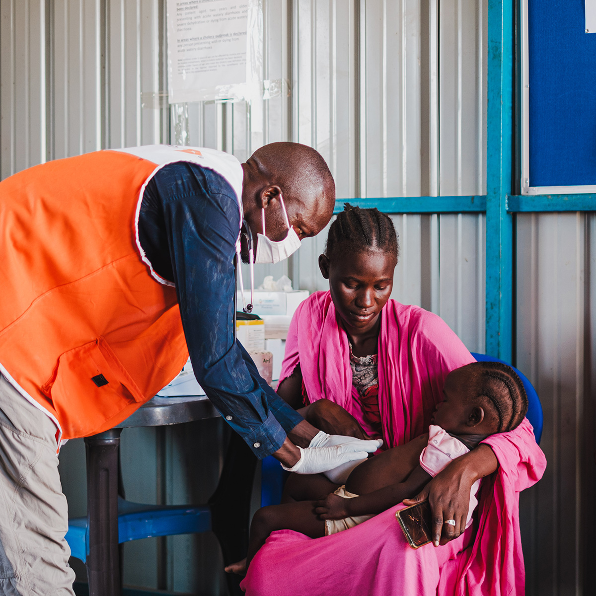 A woman in a pink dress sits on a chair in a clinic, holding her little girl as a male doctor examines her stomach.